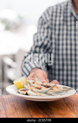 Kellner in einem karierten Hemd, der einen Teller Fisch am Strand servierte. Holztisch und selektiver Fokus. Stockfoto