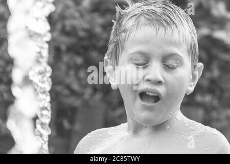 Schöne Graustufenaufnahme eines jungen kaukasischen Jungen, der mit spielt Wasser in einem Hof und Spaß haben Stockfoto