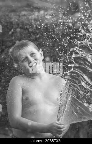 Schöne Graustufenaufnahme des jungen kaukasischen Jungen, der mit Wasser spielt In einem Hof und Spaß haben Stockfoto