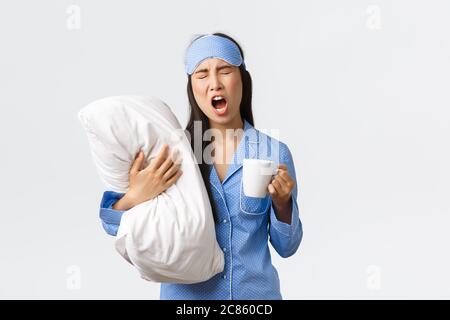 Morgen Lifestyle, Frühstück und Menschen Konzept. Mädchen mit Schlaflosigkeit in Schlafmaske und Pyjamas, umarmende Kissen, Kaffee trinken und Gähnen, versuchen Stockfoto