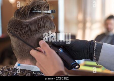 Ein Friseur schneidet einen bärtigen jungen Kerl mit einem Haarschneider und kämmt die Haare auf seinem Kopf. Die Arbeit des Meisters im Männerhaarschnitt in einem Friseurladen. Die Ba Stockfoto
