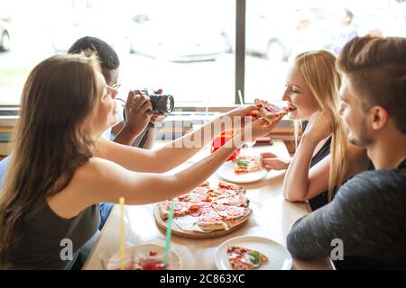 Fröhliche glückliche männliche und weibliche Studenten behandeln einander mit Bio-Pizza mit verschiedenen Belägen und nicht alchoholic Cocktails, Spaß und en