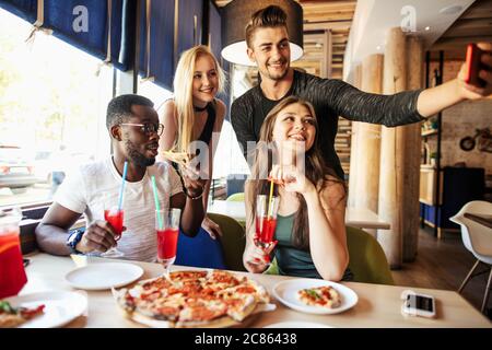 Multiethnische Gruppe von jungen fröhlichen Studenten Spaß im Café auf der Party mit leckeren Fast Food, Getränke, Selfie auf dem Smartphone Stockfoto