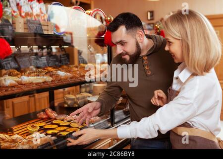 Bäckerin genießt die Arbeit in ihrer Bäckerei, hilft ihrem Kunden Dessert aus der Anzeige wählen Stockfoto