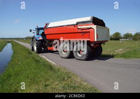 Blauer Traktor mit einem roten Anhänger, der hinter ihm gekoppelt ist, fährt auf einer Straße entlang der Wiesen in der Nähe des niederländischen Dorfes Bergen. Frühling, Mai, Niederlande Stockfoto