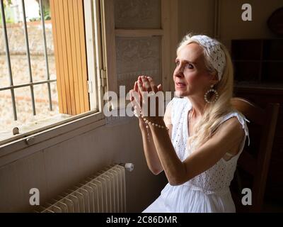 Horizontales Porträt einer reifen Frau, die mit einem Rosenkranz am Fenster betet Stockfoto