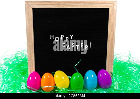 Ein schwarzes Schild mit einem Birkenrahmen, der Frohe Ostern in weißen Buchstaben mit einem Satz Regenbogen Eier und grünes Ostergras vor ihm auf einem reinen Whi sagt Stockfoto