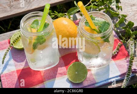 Kaltes Erfrischungsgetränk im Glasgefäß mit Zitronenscheiben und Limetten. Dekoriert mit frischen Minzkräutern und Trinkhalm auf Holzhintergrund. Stockfoto