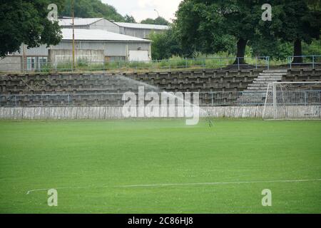 Altes Fußballstadion mit Tribüne mit verfallenen Holzsitzen oder Banken in einer Provinzstadt in Osteuropa. Der Rasen, das Spielfeld wird bewässert. Stockfoto