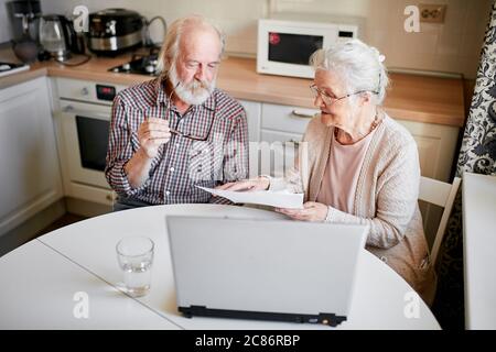 Inhalt Senior bärtig grauköpfigen Mann und seine Frau lächelnd Überprüfung Stromrechnungen oder Versicherungen am Computer mit einfachem Zugang, sitzen in der Küche ta Stockfoto