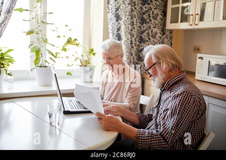 Inhalt Senior bärtig grauköpfigen Mann und seine Frau lächelnd Überprüfung Stromrechnungen oder Versicherungen am Computer mit einfachem Zugang, sitzen in der Küche ta Stockfoto