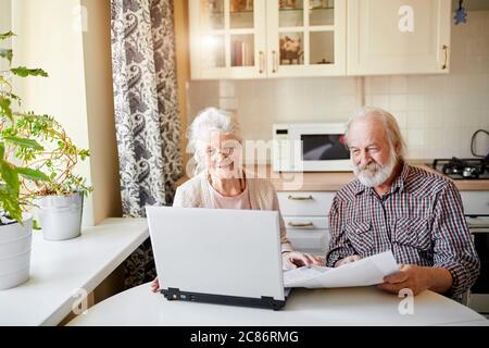 Happy Senior kaukasischen Paar tun Online-Lebensmittel einkaufen nach ihrer Liste in den Händen, die Wahl Lieblings-Dinge, zeigt auf den Bildschirm des Laptops, während Stockfoto