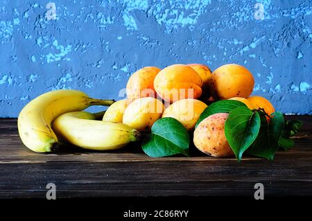 Reife Aprikosen und Bananen mit grünen Blättern auf einem Holztisch. Stockfoto