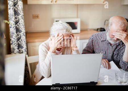 Verzweifeltes reifes Paar mit Rechnungen, die mit den Händen in der Nähe von Tempeln auf den Laptop schauen, sich nach dem Erhalt einer E-Mail mit Schuldvermerk ärgern. Grau- Stockfoto