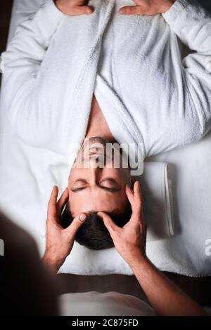 Blick aus der Höhe auf einen kaukasischen jungen Mann, der im Spa-Center eine Gesichtsmassage erhält. Professionelle Masseuse Freigabe Spannung der männlichen Kunden tun ther Stockfoto