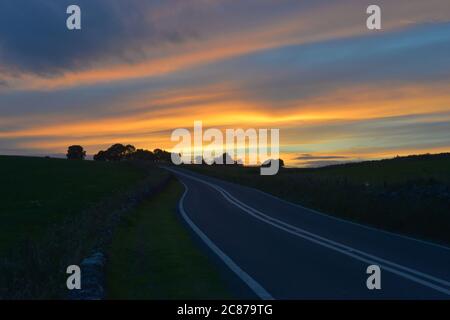 Deep Blue Frische Asphaltstraße im Peak Bezirk führt zu einigen Bäumen und einem starken orangen Sonnenuntergang. Stockfoto