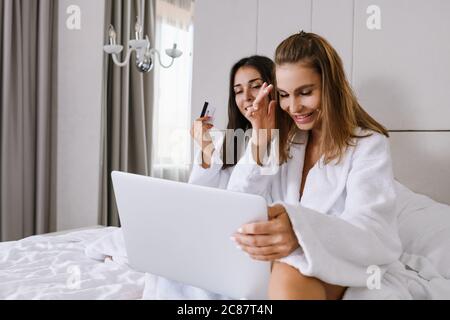 Zwei schöne lachende Frauen in weißem Bett, in Morgenbademänteln, online mit Kreditkarte und Laptop im Luxus-Appartement, Hotel Bac Stockfoto