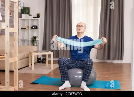 Alter Mann, der das Widerstandsband während seines Trainings benutzt. Alter Rentner online Internet-Trainingstraining zu Hause Sportliche Aktivität mit Hantel, Widerstandsband, schweizer Ball im älteren Rentenalter. Stockfoto