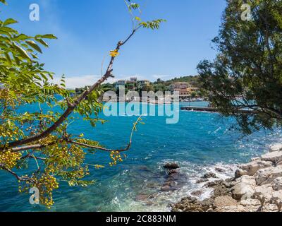 Blick auf den Hafen und bunte Häuser Dorf Kassiopi mit Bäumen, Felsen und Meer. Kassiopi ist ein touristisches Dorf im Norden der Insel Korfu Stockfoto