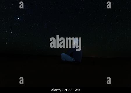 Ein einsamer Mann sitzt auf dem Wüstensand und schaut auf den Sternenhimmel, zwischen Dünen in der Wüste von Erg Chebbi. Astrofotografie Stockfoto