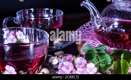 Ein Becher roten Tees und eine Teekanne in weißen Hibiskusblüten und grünen Blättern medizinischen Tees auf einem Holzständer.Zen-Tee Zeremonie. Foto von roten Kräuter Indi Stockfoto