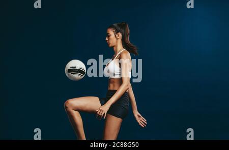 Frau Fußballspielerin jongliert einen Ball mit dem Knie vor dunklem Hintergrund. Frau in Fitness-Kleidung konzentriert sich auf Jonglieren einen Fußball. Stockfoto