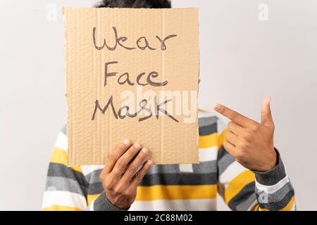 Junger Mann hält ein Pappschild, indem er sein Gesicht bedeckt, sagt, tragen Gesichtsmaske Nachricht auf isoliertem Hintergrund vor Coronavirus oder covid-19 zu schützen. Stockfoto