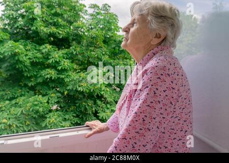 Alte ältere Frau auf dem Balkon und Blick auf Bäume Alte Dame auf der Terrasse mit Blick nach draußen. Ältere Frauen auf dem Balkon während der Quarantäne. Bleiben Stockfoto