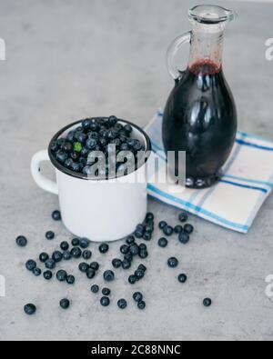 Frische Heidelbeere mit Tropfen Wasser in weißer Tasse. Heidelbeersirup in Glasflasche oder Mischung, auf Holzgrund. Stockfoto