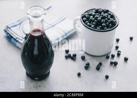 Frische Heidelbeere mit Tropfen Wasser in weißer Tasse. Heidelbeersirup in Glasflasche oder Mischung, auf Holzgrund. Stockfoto
