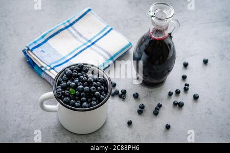 Frische Heidelbeere mit Tropfen Wasser in weißer Tasse. Heidelbeersirup in Glasflasche oder Mischung, auf Holzgrund. Stockfoto