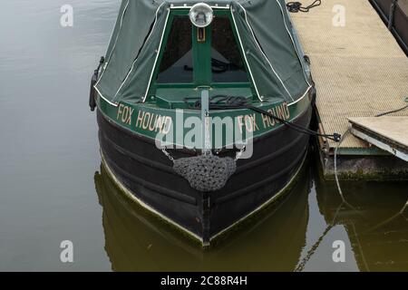 Vorderansicht eines gut gepflegten schmalen Bootes gesehen, das in seinem Binnenschiffhafen festgemacht ist. Ein Gehweg kann gesehen werden, um Zugang zum Boot zu erhalten. Stockfoto