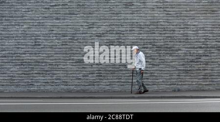 Ein älterer Mann mit Stock geht an einer Steinmauer entlang Stockfoto