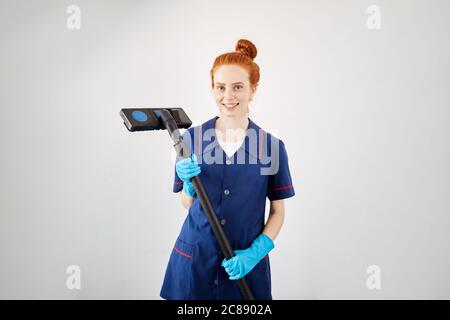 Kaukasische Haushälterin mit rotem Haargummi, blauem Arbeitsmantel und Gummihandschuhen, die die Kamera mit der Bürste betrachten. Hausarbeit, Vakuum cl Stockfoto