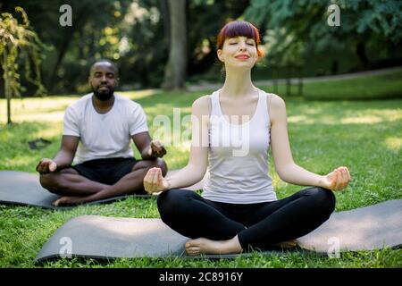 Konzept von Yoga und Meditation, Familienleben. Zwei junge multiethnische Freunde, kaukasische Frau und afrikanischer Mann, sitzen in Lotuspose auf Yogamatten Stockfoto