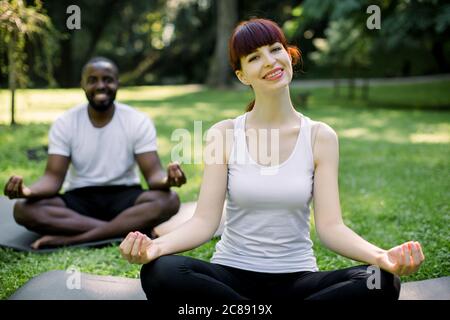 Vorderansicht des multiethnischen Paares, das im Park Yoga praktiziert. Lächelnd hübsche kaukasische Frau, die in Lotushaltung auf der Matte sitzt. Fröhlicher afrikanischer Mann Stockfoto