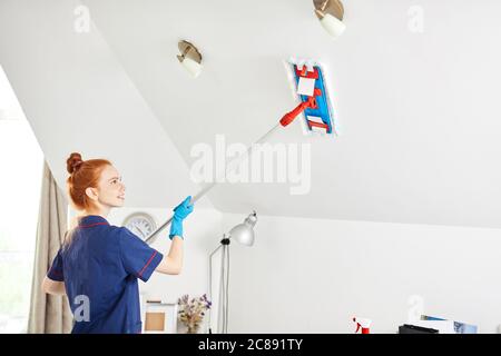 Schöne rothaarige junge Frau in blauem Arbeitsoutfit mit einem Mopp beim Reinigen der weißen Decke im weißen Raum. Stockfoto