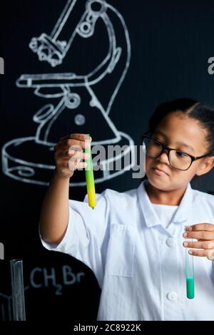 Mischrasse asiatische, afrikanische Mädchen in Schutzbrille tragen weiße Labor hält Reagenzgläser und Kolben Durchführung von Experimenten über schwarze Tafel mit symb Stockfoto