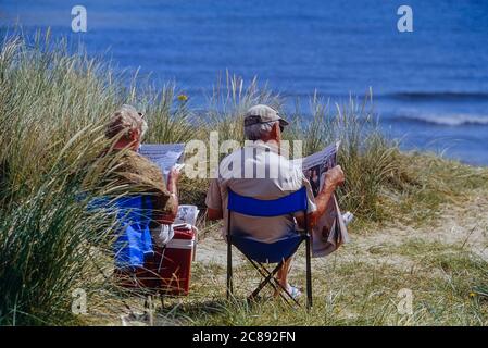 Ein älteres Paar sitzt am Strand und liest die Zeitungen. Hemsby, Great Yarmouth, East Anglia, England. GROSSBRITANNIEN Stockfoto