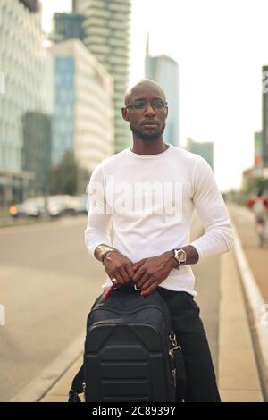 Junger stilvoller afrikanischer Mann mit Brille, die einen Rucksack hält Die Straße unter dem Sonnenlicht Stockfoto