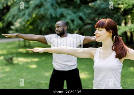 Konzept für einen gesunden Familienleben und Yoga im Park. Junge multiethnische Paar trainieren im Freien mit ausgestreckten Armen, Blick nach vorne. Konzentrieren Sie sich auf Stockfoto