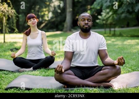 Nahaufnahme eines multirassischen Paares, das sich im Park ausruhen lässt, Yoga praktiziert, in Lotusposition auf Yogamatten sitzt, Hände im Zen, tiefes Atmen Stockfoto