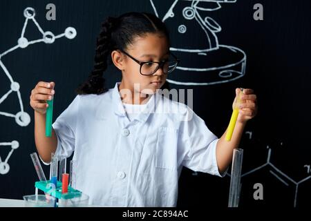 Mischrasse asiatische, afrikanische Mädchen in Schutzbrille tragen weiße Labor hält Reagenzgläser und Kolben Durchführung von Experimenten über schwarze Tafel mit symb Stockfoto