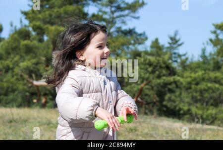 Ein kleines lächelndes weißes Mädchen mit langen Haaren hält einen Roller-Griff in ihren Händen.EIN Kind in warmen Kleidern fährt einen Roller im Wald. Stockfoto