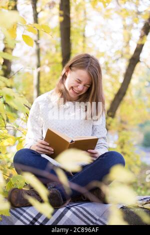 Schöne Teenager Mädchen sitzt im Herbstgarten am Zaun, auf Wolldecken karierten und lachend Lesen lustige Buch, Zahnspange Stockfoto