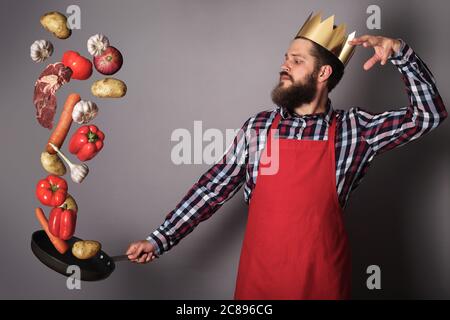 Kochen man Concept, König von Küche, bärtigen Mann in kariertes Hemd, Nach oben Drop Fleisch und Gemüse aus der Pfanne, Studio shot auf grauem Hintergrund Stockfoto