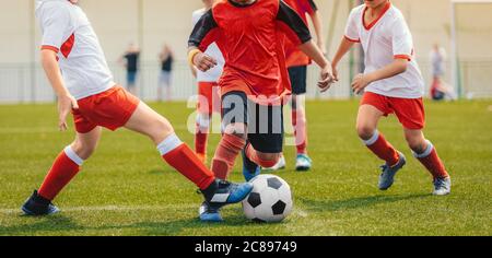 Multiethnische Kinder spielen Fußballspiel. Junge Jungs laufen nach Fußball auf Gras Fußballplatz. Kinder in rot-weißen Trikots Stockfoto