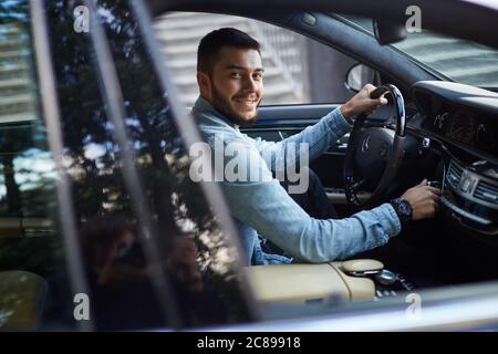 Ein schöner lächelnder Mann gekleidet lässig suchen aus einem Autofenster. Seitenansicht Foto. Let's go. Bewegung Konzept Stockfoto