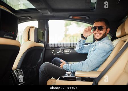 Fröhlicher junger Mann in Jeansjacke sitzt auf dem Auto und posiert vor der Kamera. Seitenansicht Foto. Stockfoto
