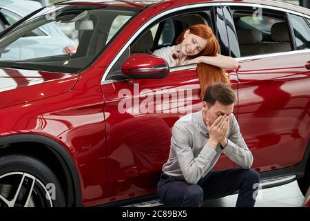 Happy Content Rothaarige Frau sitzt auf dem Fahrersitz in Luxus-Auto im Auto Showroom, Blick auf sich selbst an Seite mirrow, während ihr trauriger Freund sitzt in th Stockfoto